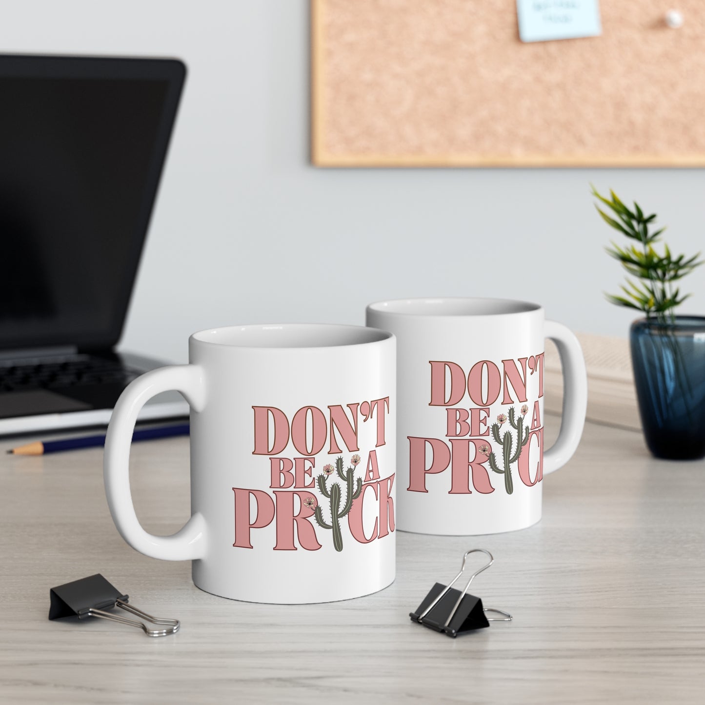 Two white ceramic mugs on a desk that say Don't Be A Prick.