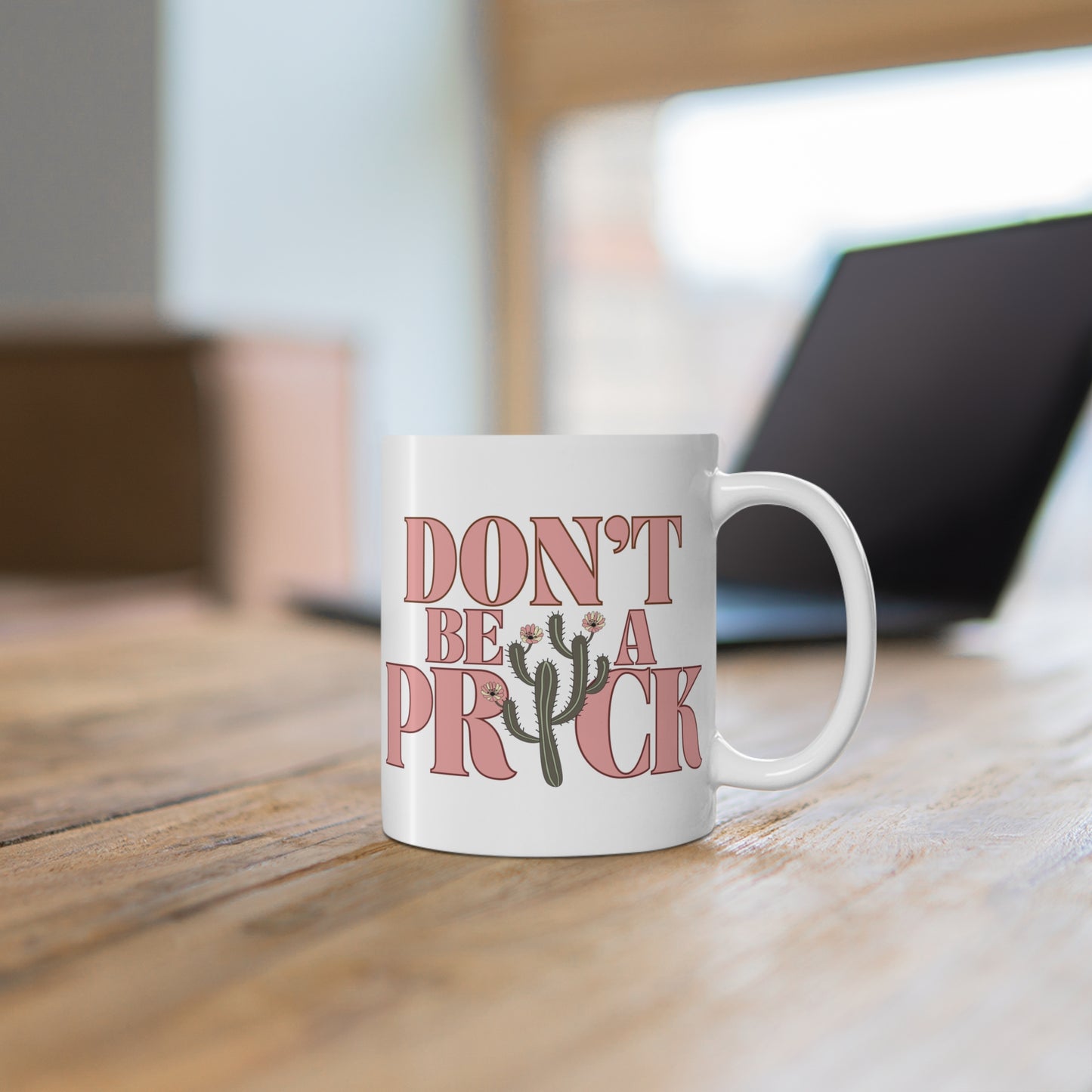 A white ceramic mug on a desk that says Don't Be A Prick.