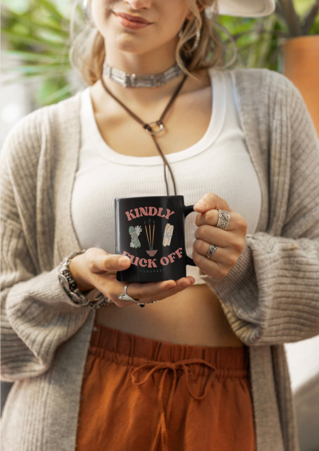 Woman holding a black mug that says Kindly Fuck Off with incense, palo santo, and sage.